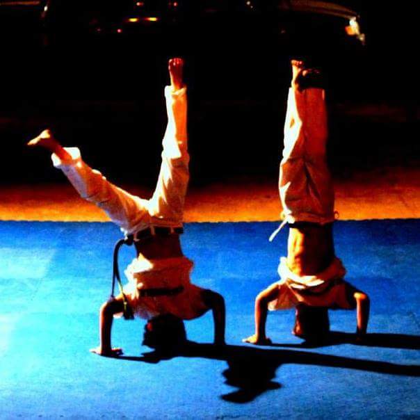 Two children in white uniforms doing headstands side by side on a blue mat