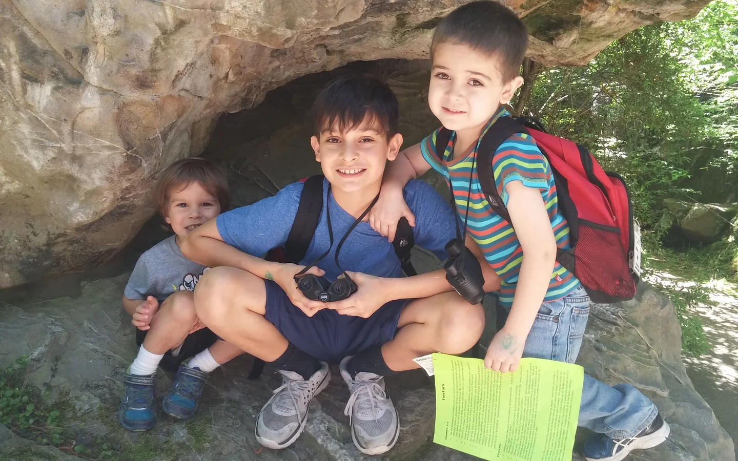 Three children resting under a rock overhang on a nature walk, with binoculars and backpacks