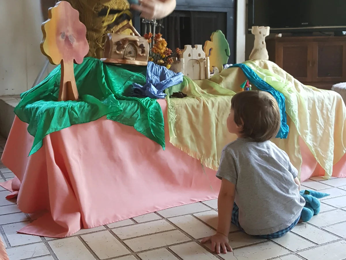 A child sitting on the floor, watching a puppet story unfold on a silk-draped table